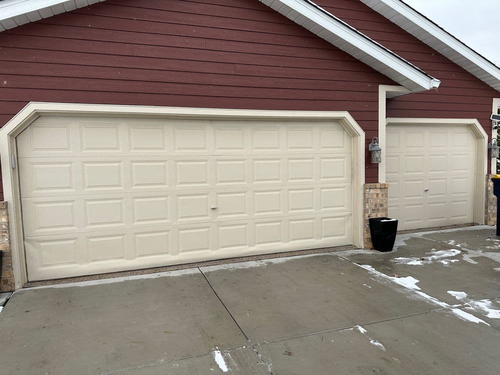 two beige garage doors on a red house with a concrete driveway and snow patches