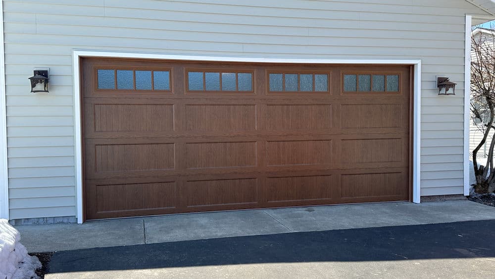 Wooden garage door with windows, installed on a house's exterior, beside decorative wall sconces.