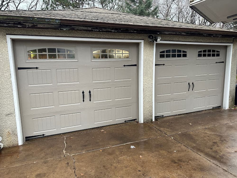 Dual beige garage doors with decorative windows and black hardware on a concrete driveway.