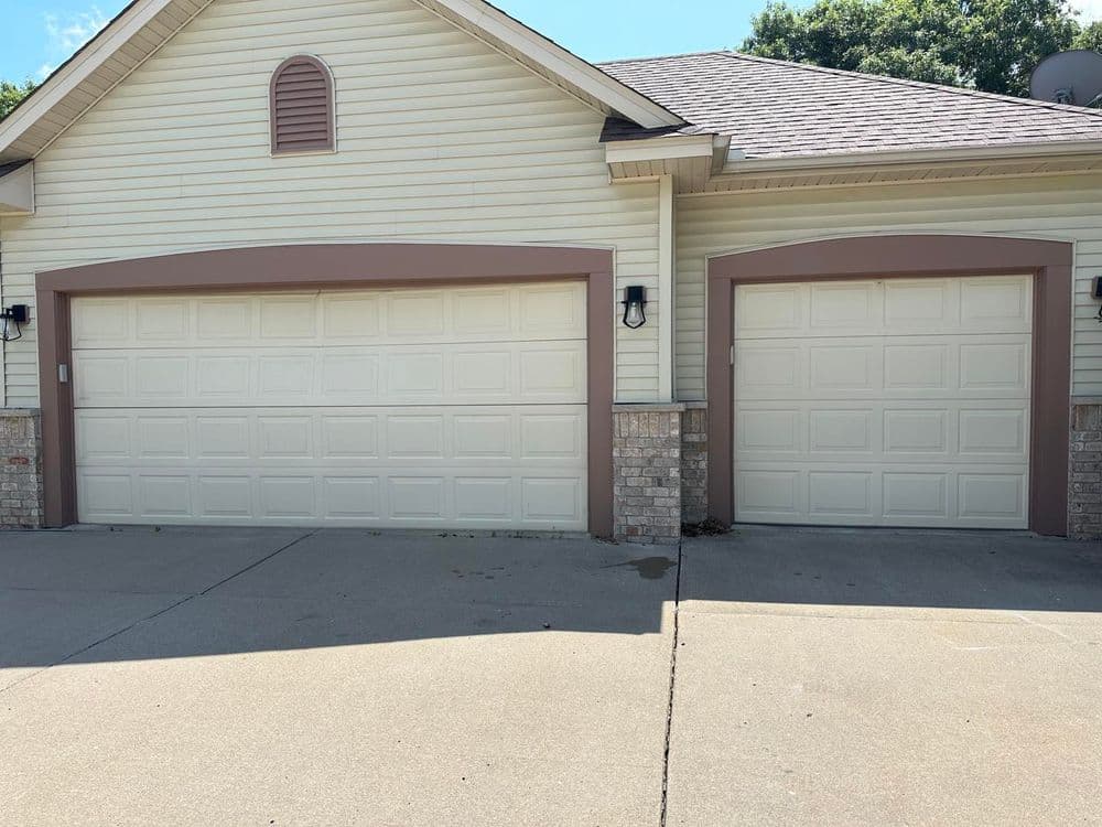 Two beige garage doors with decorative trim on a light-colored house. Driveway visible.