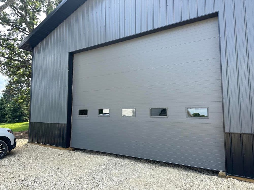 Modern gray metal garage door with windows, surrounded by gravel and greenery.