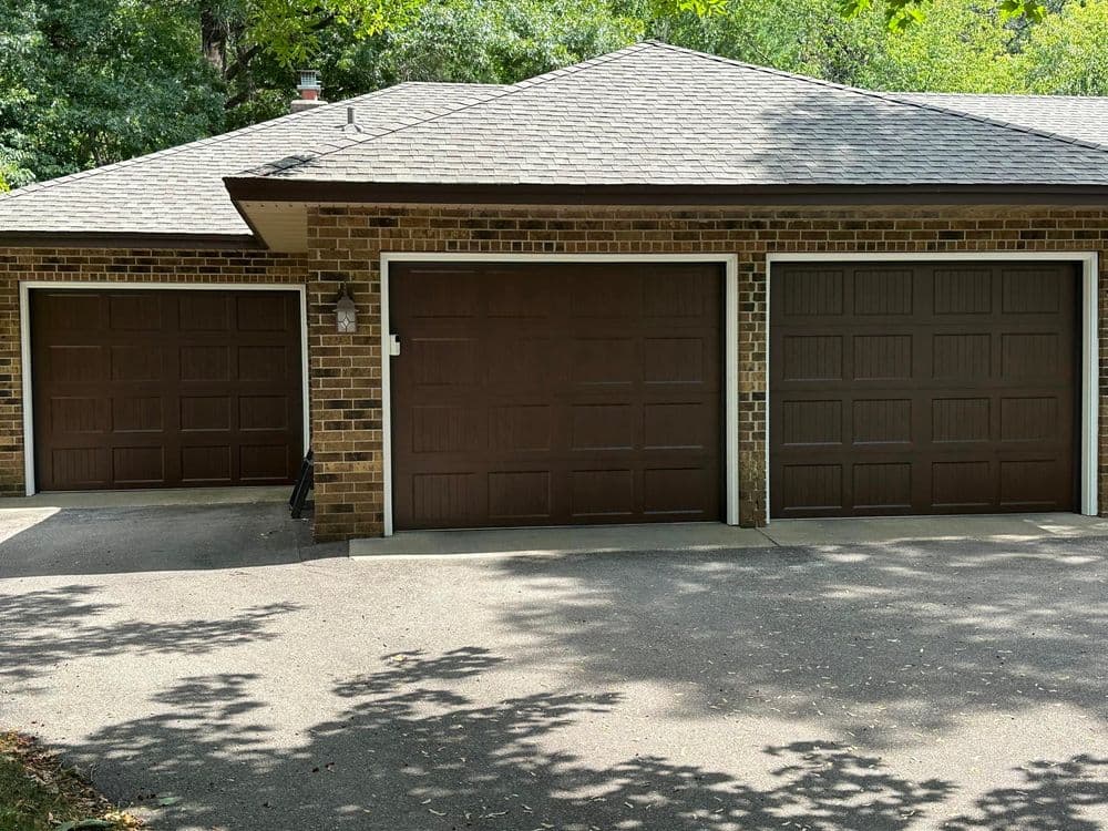 Brown garage doors on a brick house with a paved driveway and surrounding greenery.