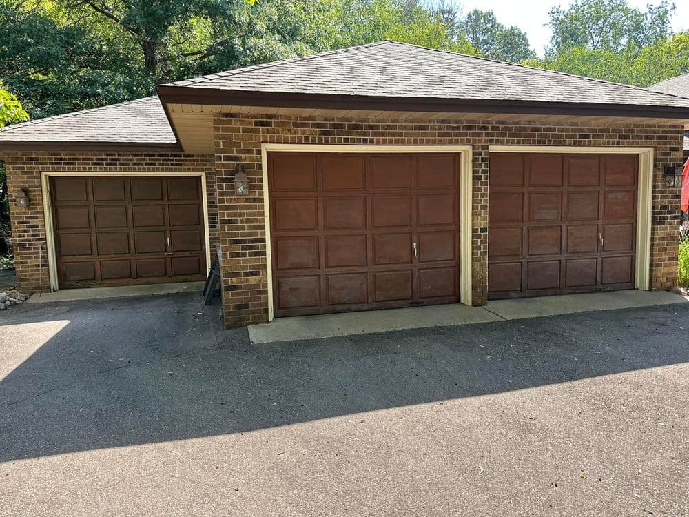 Three brown garage doors on a brick house with a paved driveway and trees in the background.
