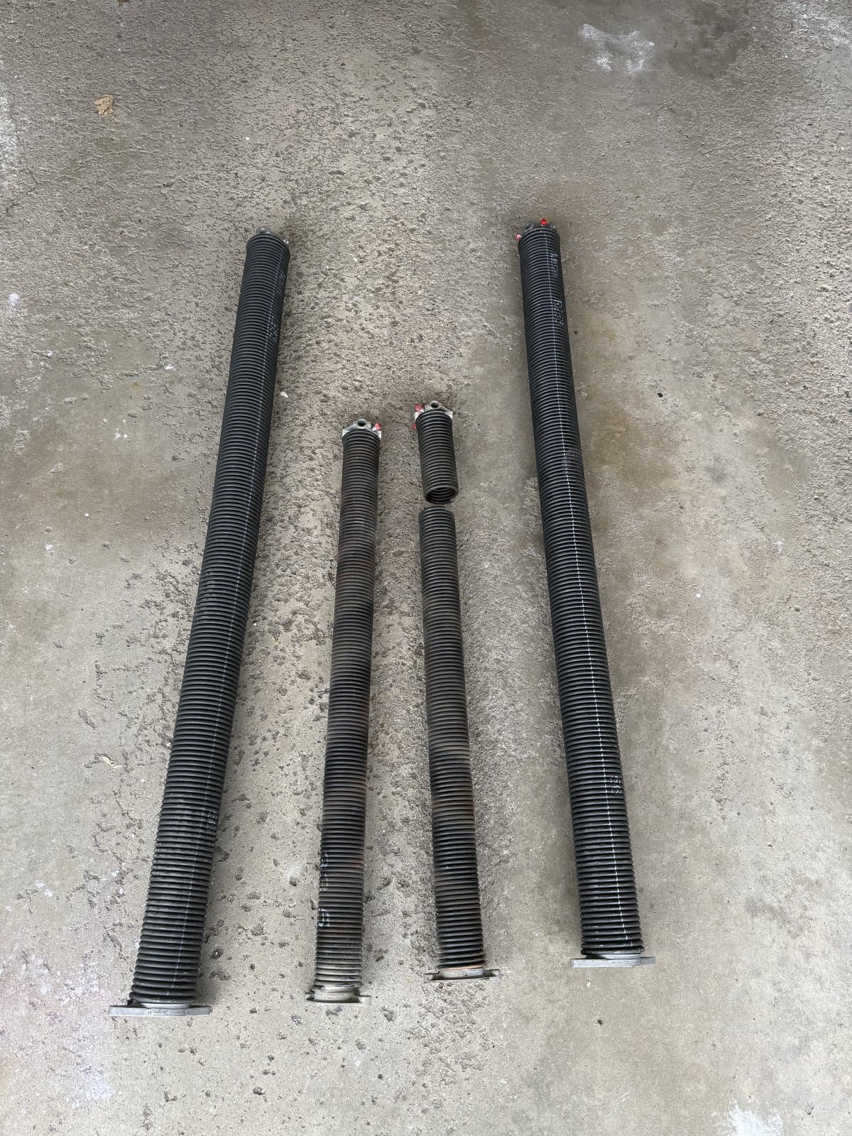 A broken pair of standard garage door springs next to a new pair of high-cycle torsion springs on a garage floor